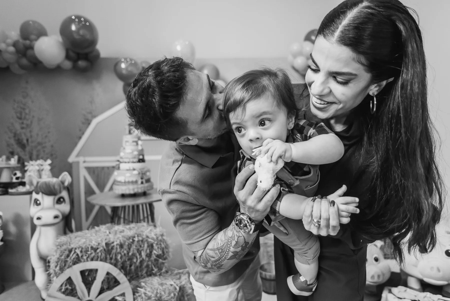 Pai beijando o bebê no colo da mãe, numa festa de aniversário — fotografia em preto e branco.